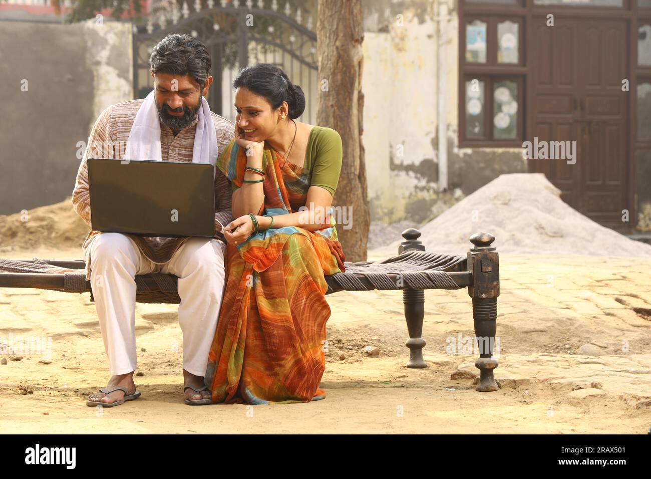 Heureuse famille indienne rurale dans le village. Couple rural assis ensemble souriant sur lit bébé devant leur maison dans la cour avant en utilisant un ordinateur portable pour les paiements en ligne. Banque D'Images