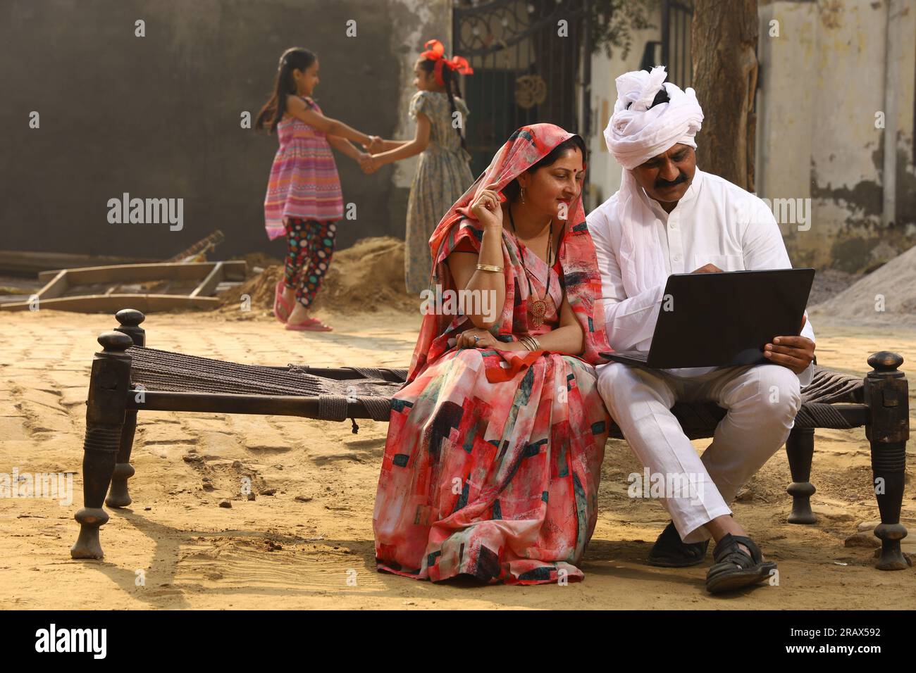 Heureuse famille indienne rurale dans le village. Couple rural assis ensemble souriant sur lit bébé devant leur maison dans la cour avant en utilisant un ordinateur portable pour les paiements en ligne. Banque D'Images
