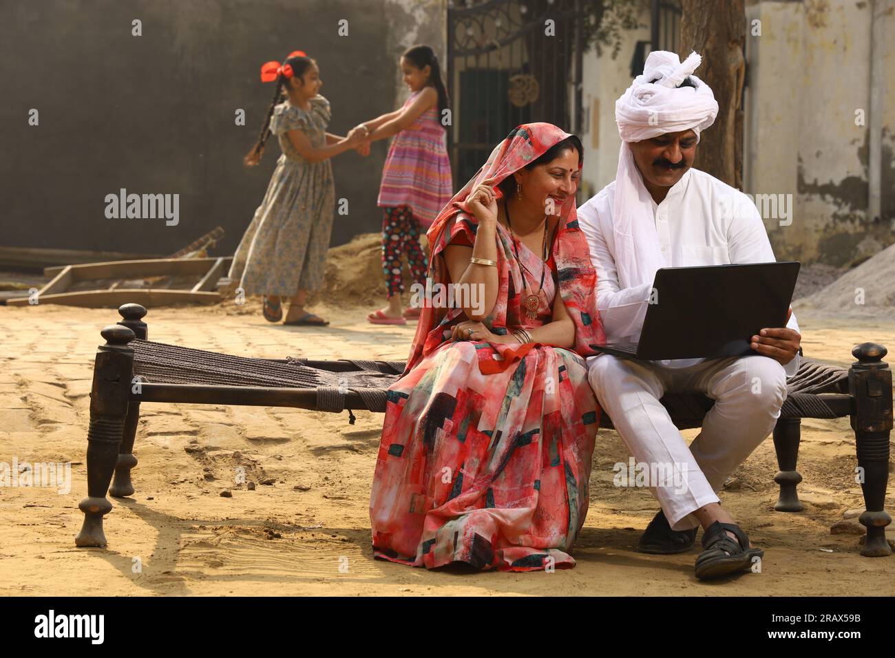 Heureuse famille indienne rurale dans le village. Couple rural assis ensemble souriant sur lit bébé devant leur maison dans la cour avant en utilisant un ordinateur portable pour les paiements en ligne. Banque D'Images