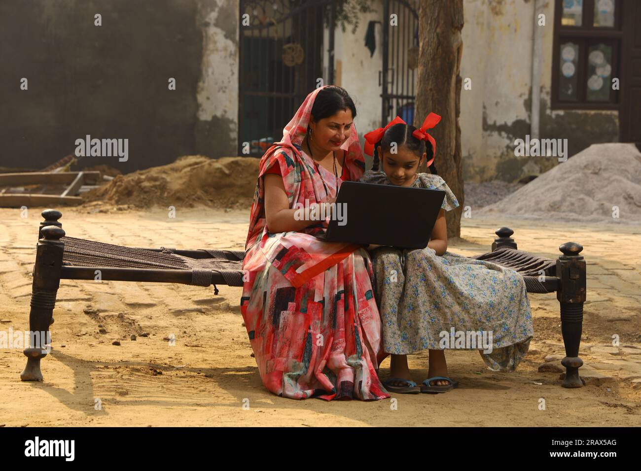 Heureuse famille indienne rurale dans le village. Couple rural assis ensemble souriant sur lit bébé devant leur maison dans la cour avant en utilisant un ordinateur portable pour les paiements en ligne. Banque D'Images