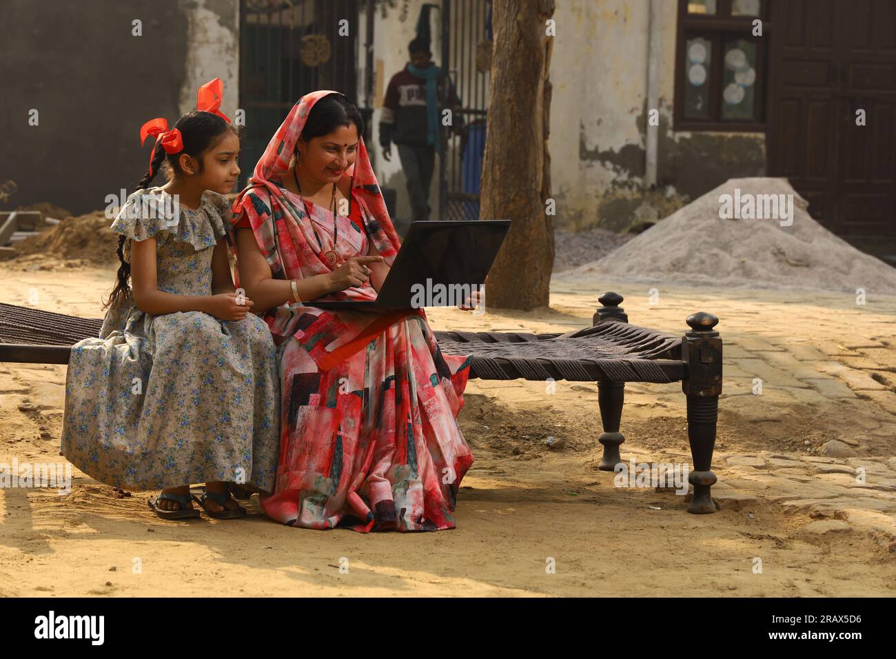 Heureuse famille indienne rurale dans le village. mère portant un saree traditionnel et sa fille assis ensemble sur un lit bébé devant leur maison dans la cour avant. Banque D'Images