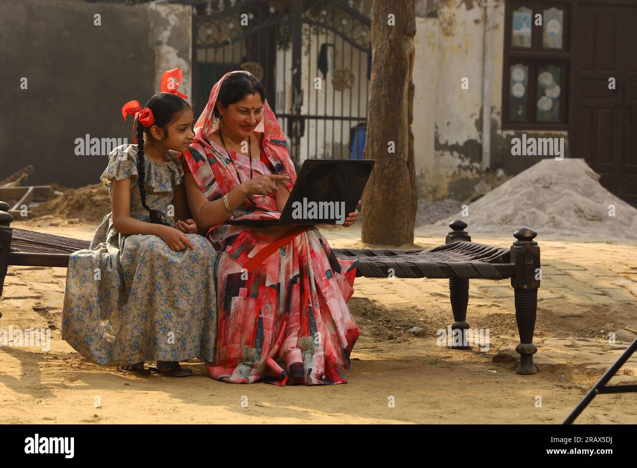 Heureuse famille indienne rurale dans le village. mère portant un saree traditionnel et sa fille assis ensemble sur un lit bébé devant leur maison dans la cour avant. Banque D'Images