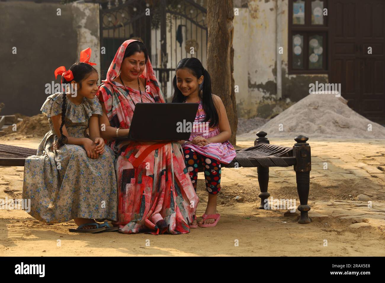 Heureuse famille indienne rurale dans le village. mère portant un saree traditionnel et sa fille assis ensemble sur un lit bébé devant leur maison dans la cour avant. Banque D'Images