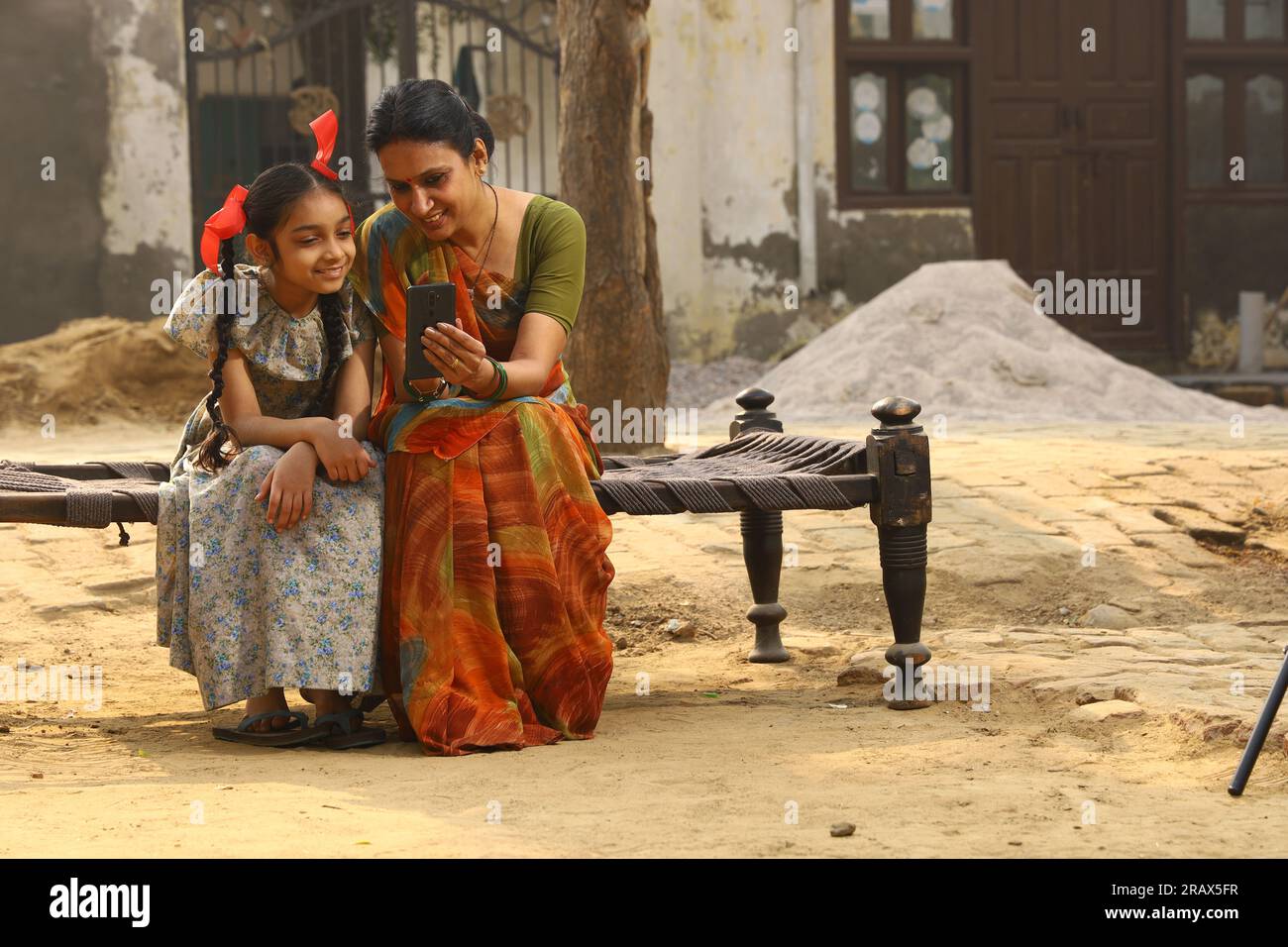 Heureuse famille indienne rurale dans le village. Mère et fille assis ensemble souriant sur un lit bébé devant leur maison dans la cour avant en utilisant un téléphone portable. Banque D'Images