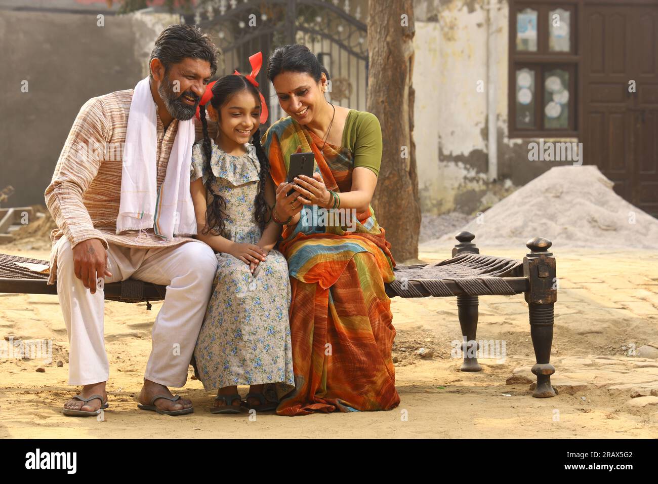 Heureux Indien famille rurale de mère, père et fille assis ensemble souriant sur lit à l'extérieur de leur maison dans la cour avant en utilisant le téléphone portable. Banque D'Images