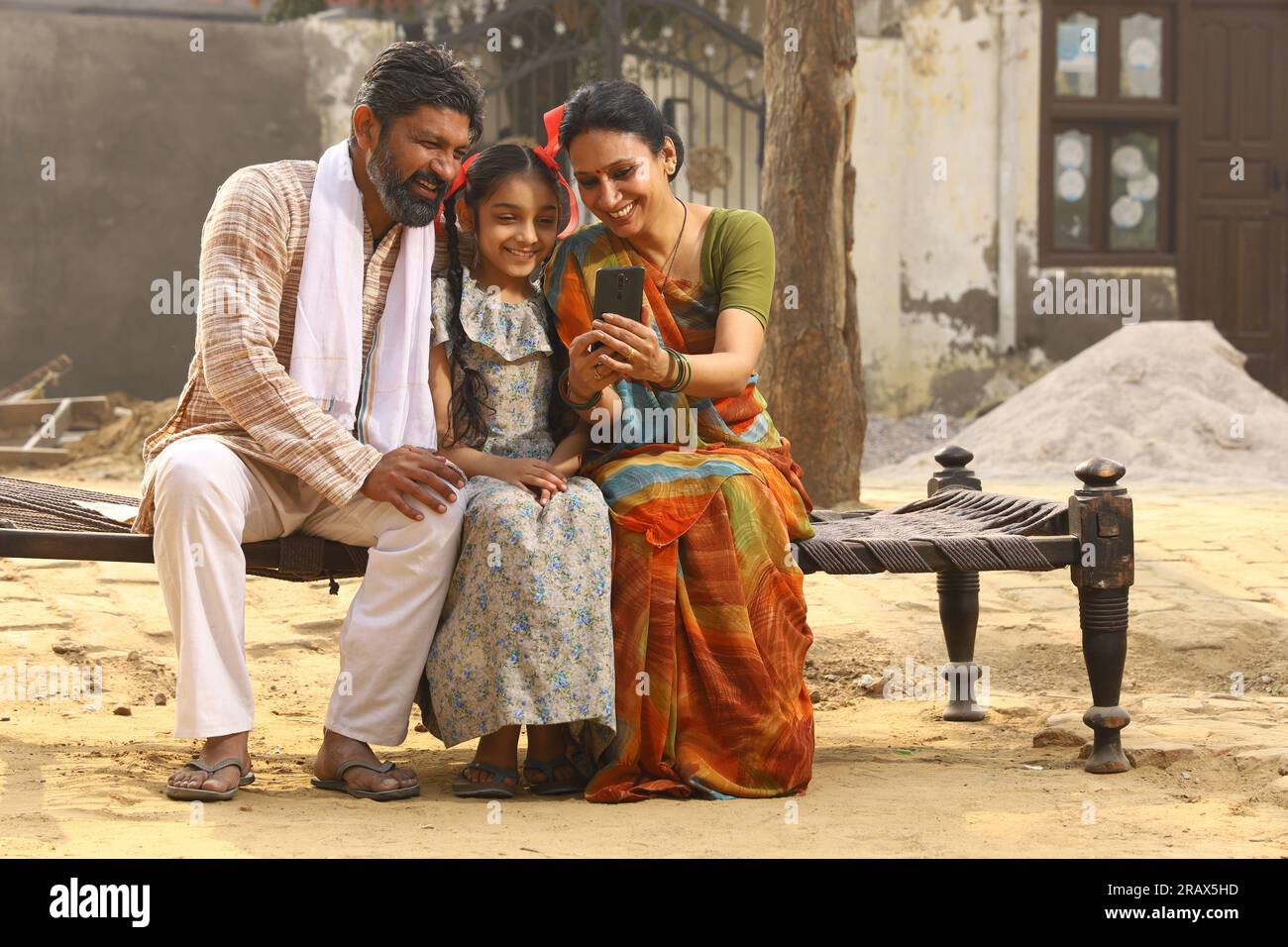 Heureux Indien famille rurale de mère, père et fille assis ensemble souriant sur lit à l'extérieur de leur maison dans la cour avant en utilisant le téléphone portable. Banque D'Images