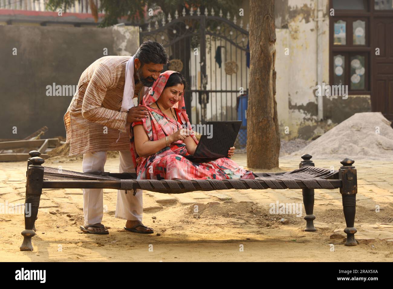 Heureuse famille indienne rurale dans le village. Couple rural assis ensemble souriant sur lit bébé devant leur maison dans la cour avant en utilisant un ordinateur portable pour les paiements en ligne. Banque D'Images