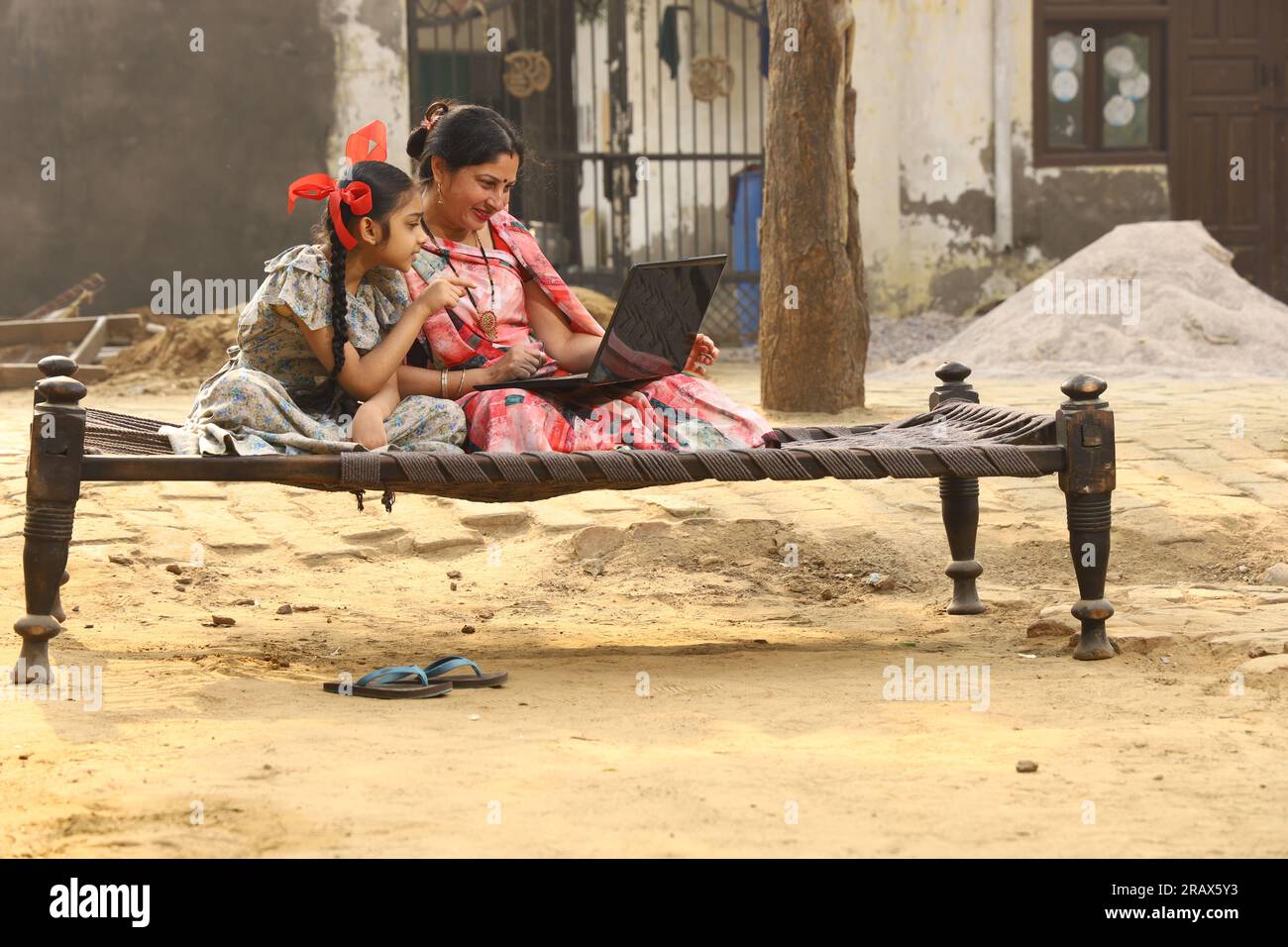 Heureuse famille indienne rurale dans le village. mère portant un saree traditionnel et sa fille assis ensemble sur un lit bébé devant leur maison dans la cour avant. Banque D'Images