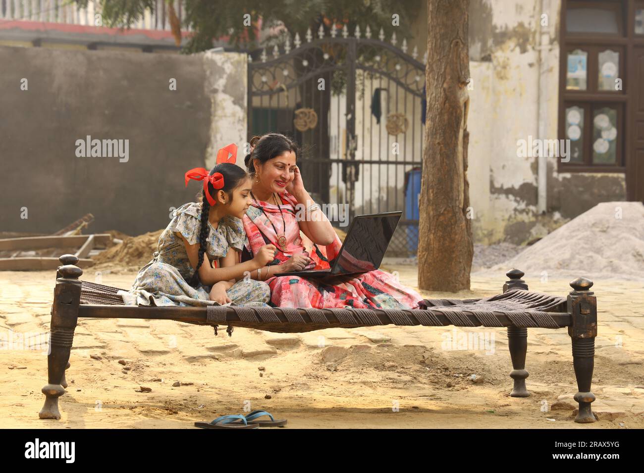 Heureuse famille indienne rurale dans le village. mère portant un saree traditionnel et sa fille assis ensemble sur un lit bébé devant leur maison dans la cour avant. Banque D'Images
