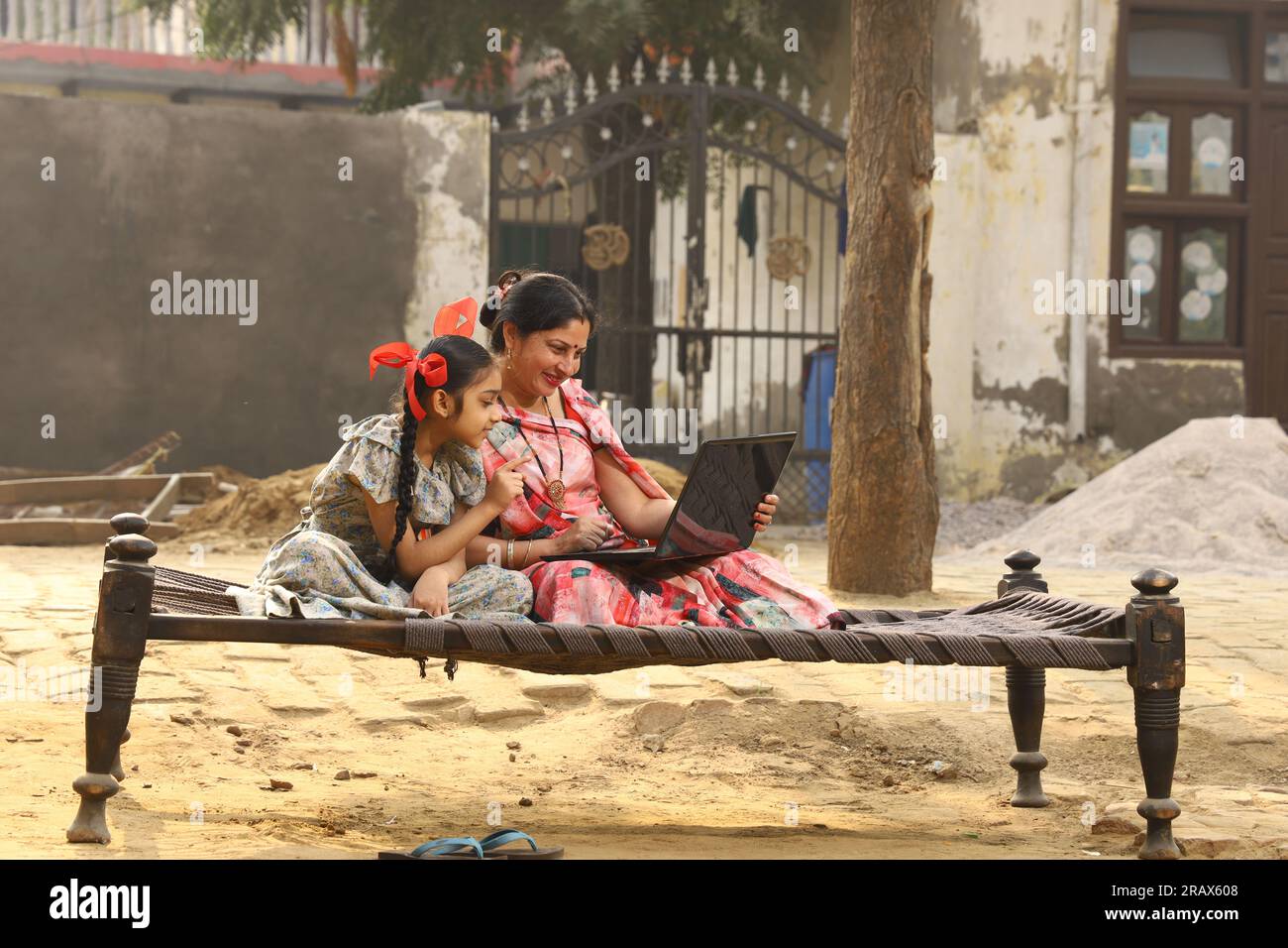 Heureuse famille indienne rurale dans le village. mère portant un saree traditionnel et sa fille assis ensemble sur un lit bébé devant leur maison dans la cour avant. Banque D'Images