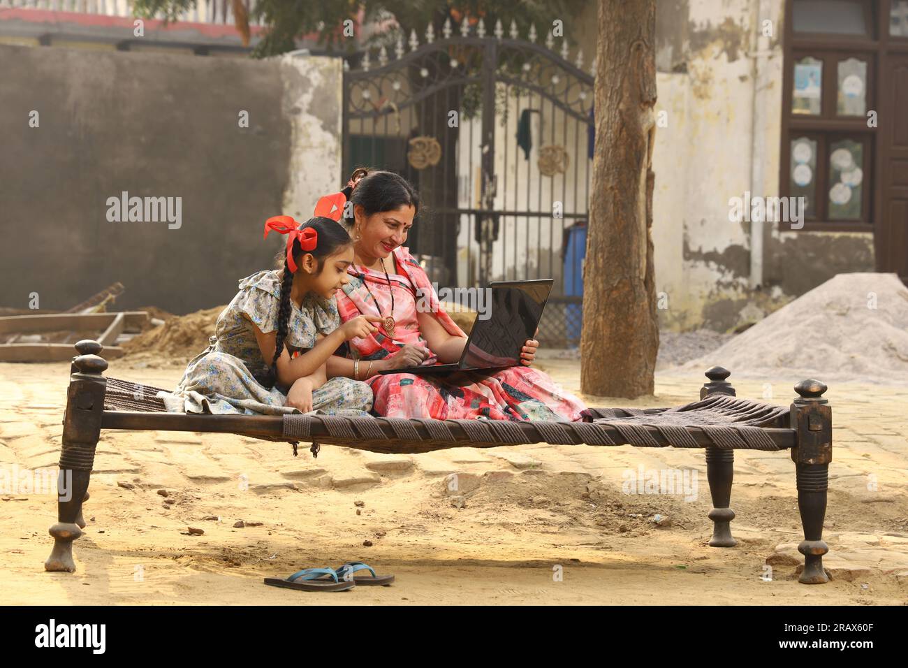 Heureuse famille indienne rurale dans le village. mère portant un saree traditionnel et sa fille assis ensemble sur un lit bébé devant leur maison dans la cour avant. Banque D'Images
