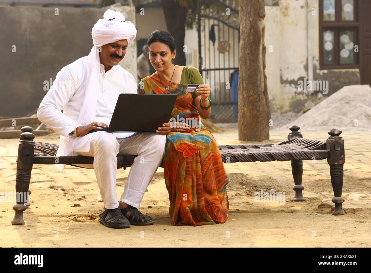 Heureuse famille indienne rurale dans le village. Couple rural assis ensemble souriant sur lit bébé devant leur maison dans la cour avant en utilisant un ordinateur portable pour les paiements en ligne. Banque D'Images