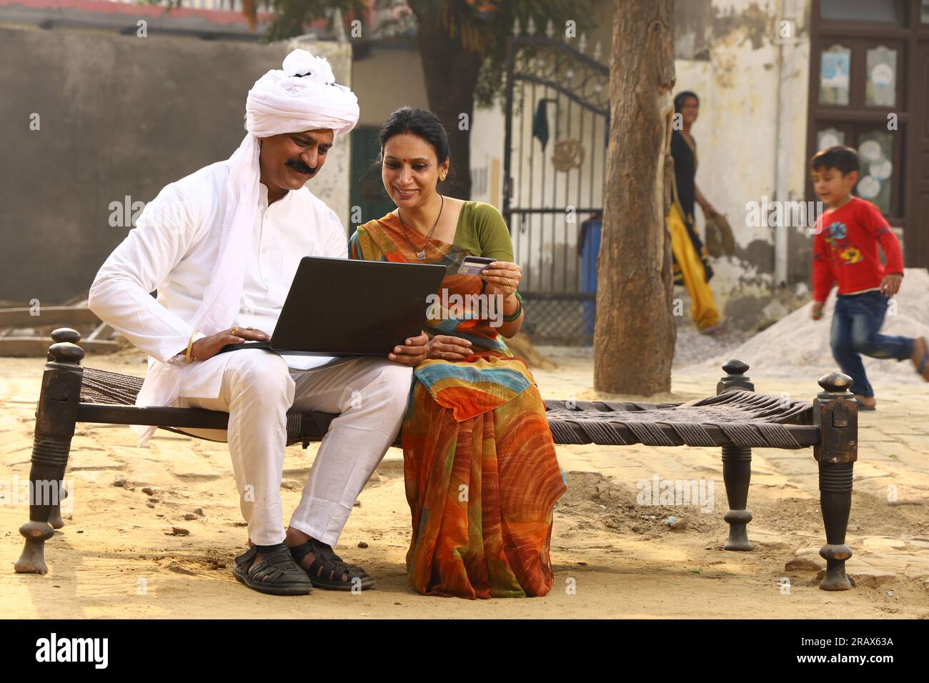 Heureuse famille indienne rurale dans le village. Couple rural assis ensemble souriant sur lit bébé devant leur maison dans la cour avant en utilisant un ordinateur portable pour les paiements en ligne. Banque D'Images