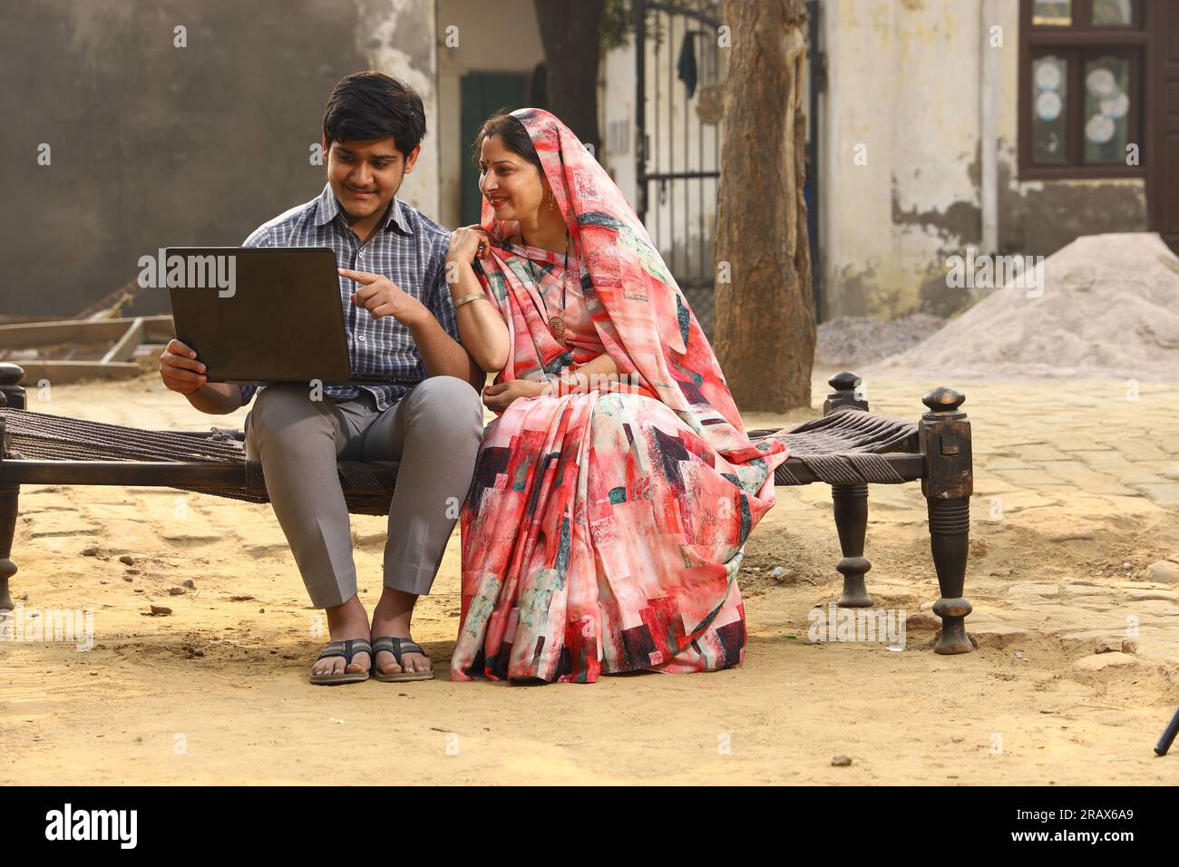 Heureux Indien Rural mère et fils assis sur un lit dans la cour avant. Mère dans belle Saree. Travailler sur un ordinateur portable. Banque D'Images