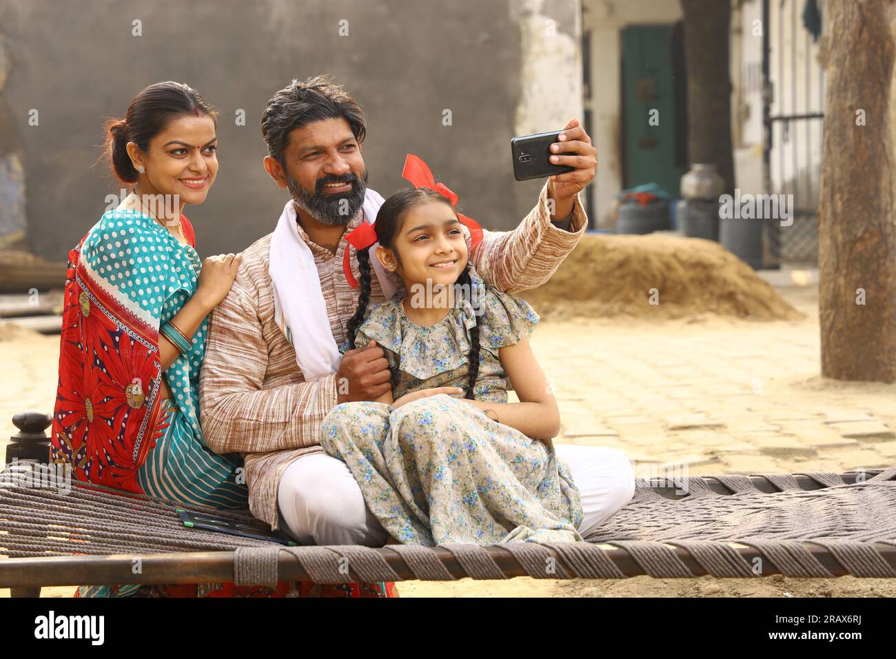 Heureux Indien famille rurale de mère, père et fille assis ensemble souriant sur lit à l'extérieur de leur maison dans la cour avant en utilisant le téléphone portable. Banque D'Images