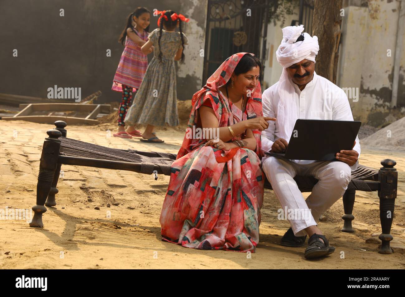 Heureuse famille indienne rurale dans le village. Couple rural assis ensemble souriant sur lit bébé devant leur maison dans la cour avant en utilisant un ordinateur portable pour les paiements en ligne. Banque D'Images