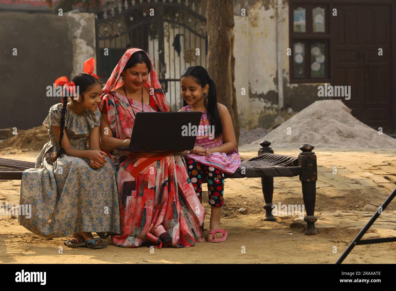 Heureuse famille indienne rurale dans le village. Mère et filles assises ensemble souriant sur un lit bébé devant leur maison dans la cour avant. Mère dans belle Saree Banque D'Images