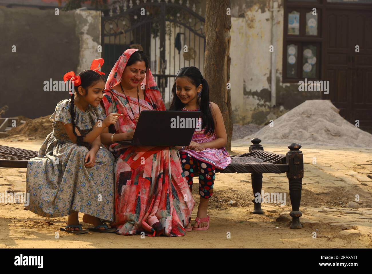 Heureuse famille indienne rurale dans le village. Mère et filles assises ensemble souriant sur un lit bébé devant leur maison dans la cour avant. Mère dans belle Saree Banque D'Images