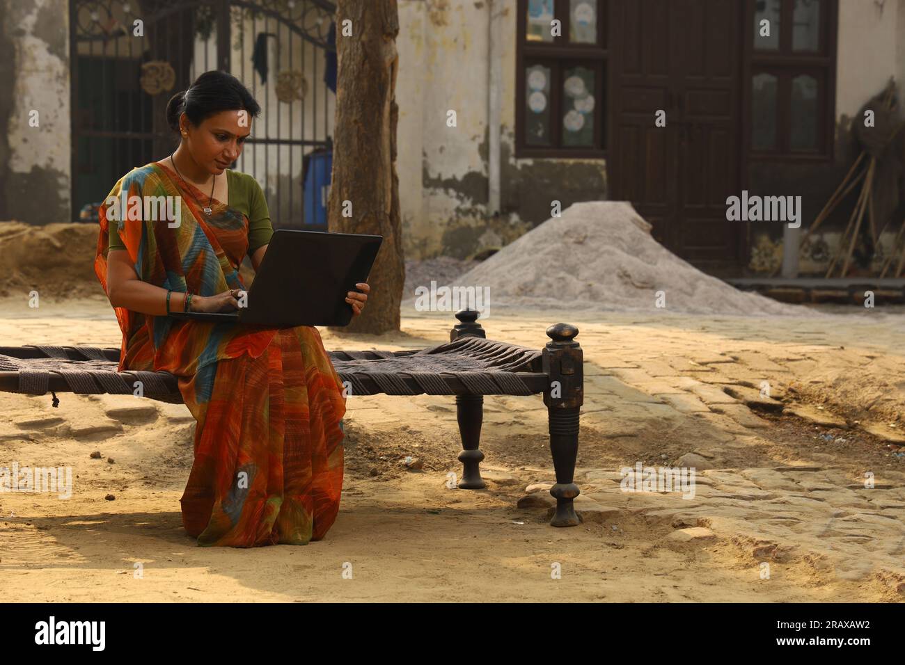 Heureuse femme rurale indienne dans le village. elle est assise sur le lit souriant dehors dans belle saree. Culture indienne, robe traditionnelle, surf sur un ordinateur portable Banque D'Images