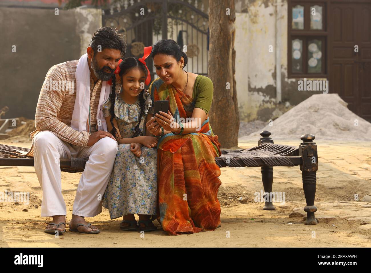 Heureux Indien famille rurale de mère, père et fille assis ensemble souriant sur lit à l'extérieur de leur maison dans la cour avant en utilisant le téléphone portable. Banque D'Images