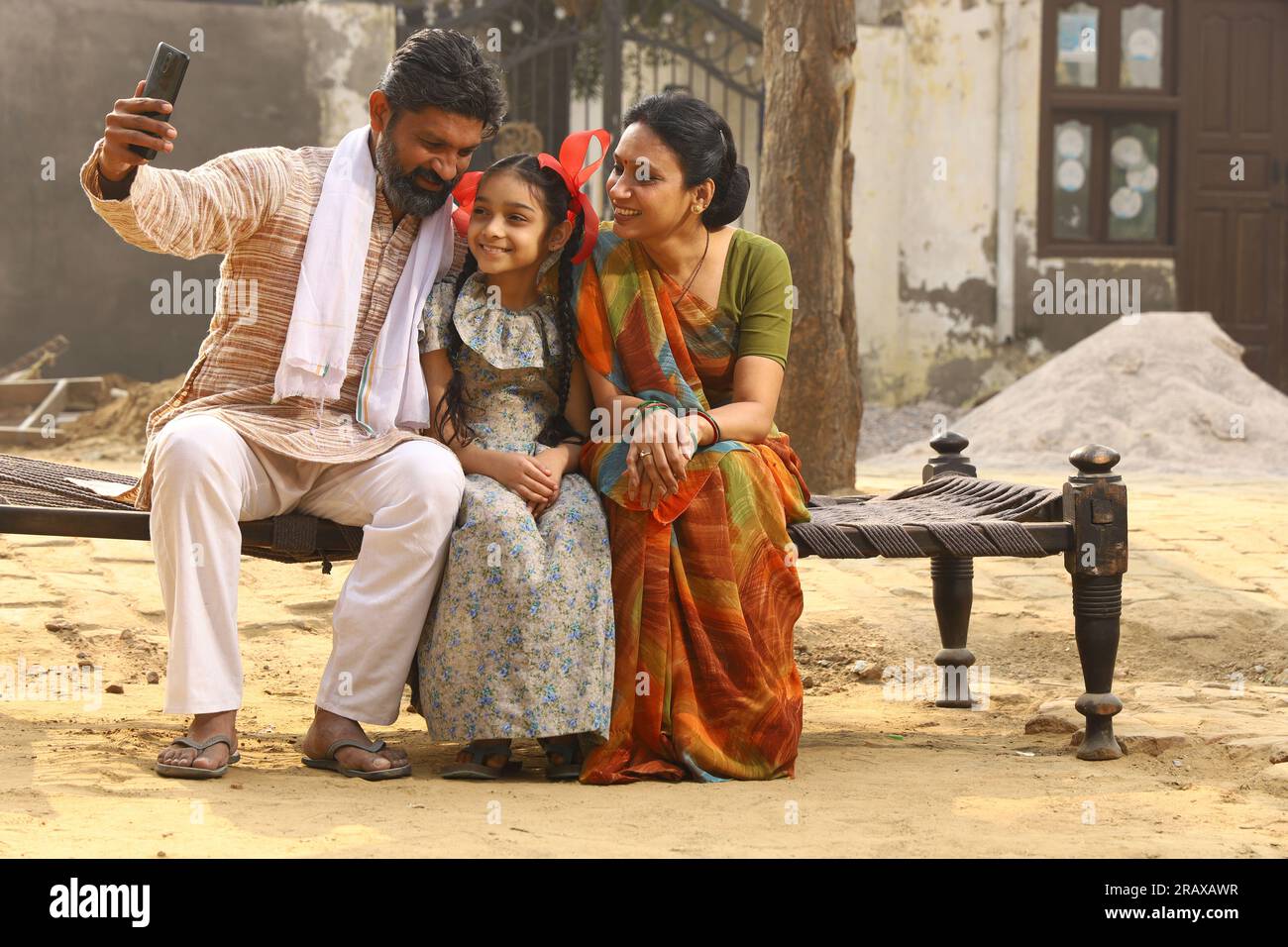 Heureux Indien famille rurale de mère, père et fille assis ensemble souriant sur lit à l'extérieur de leur maison dans la cour avant en utilisant le téléphone portable. Banque D'Images