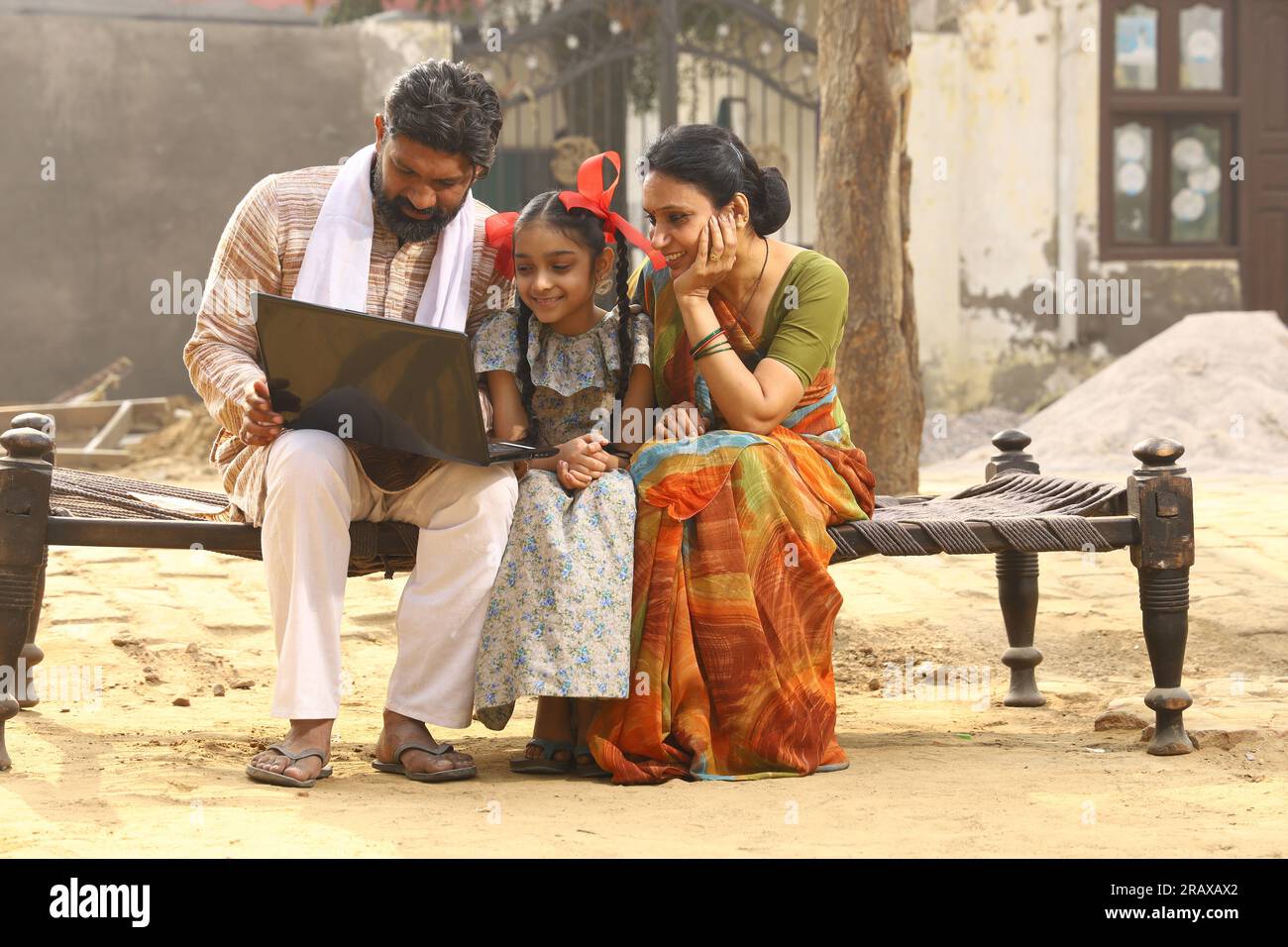 Heureux Indien famille rurale de mère, père, fille assis sur lit en dehors de leur maison dans la cour avant en utilisant ordinateur portable. pyjama sari kurta traditionnel Banque D'Images