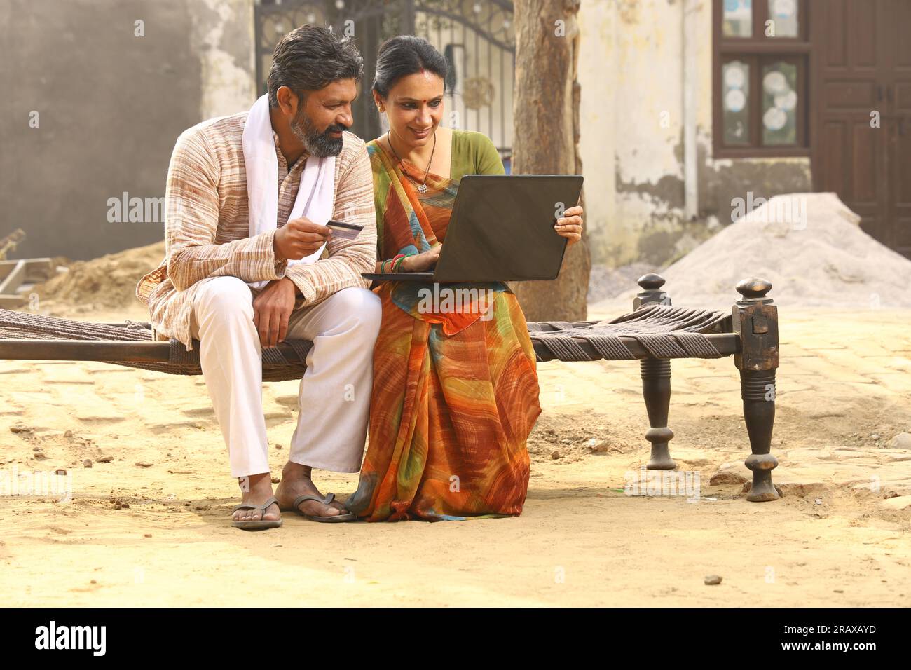 Heureuse famille indienne rurale dans le village. Couple rural assis ensemble souriant sur lit bébé devant leur maison dans la cour avant en utilisant un ordinateur portable pour les paiements en ligne. Banque D'Images