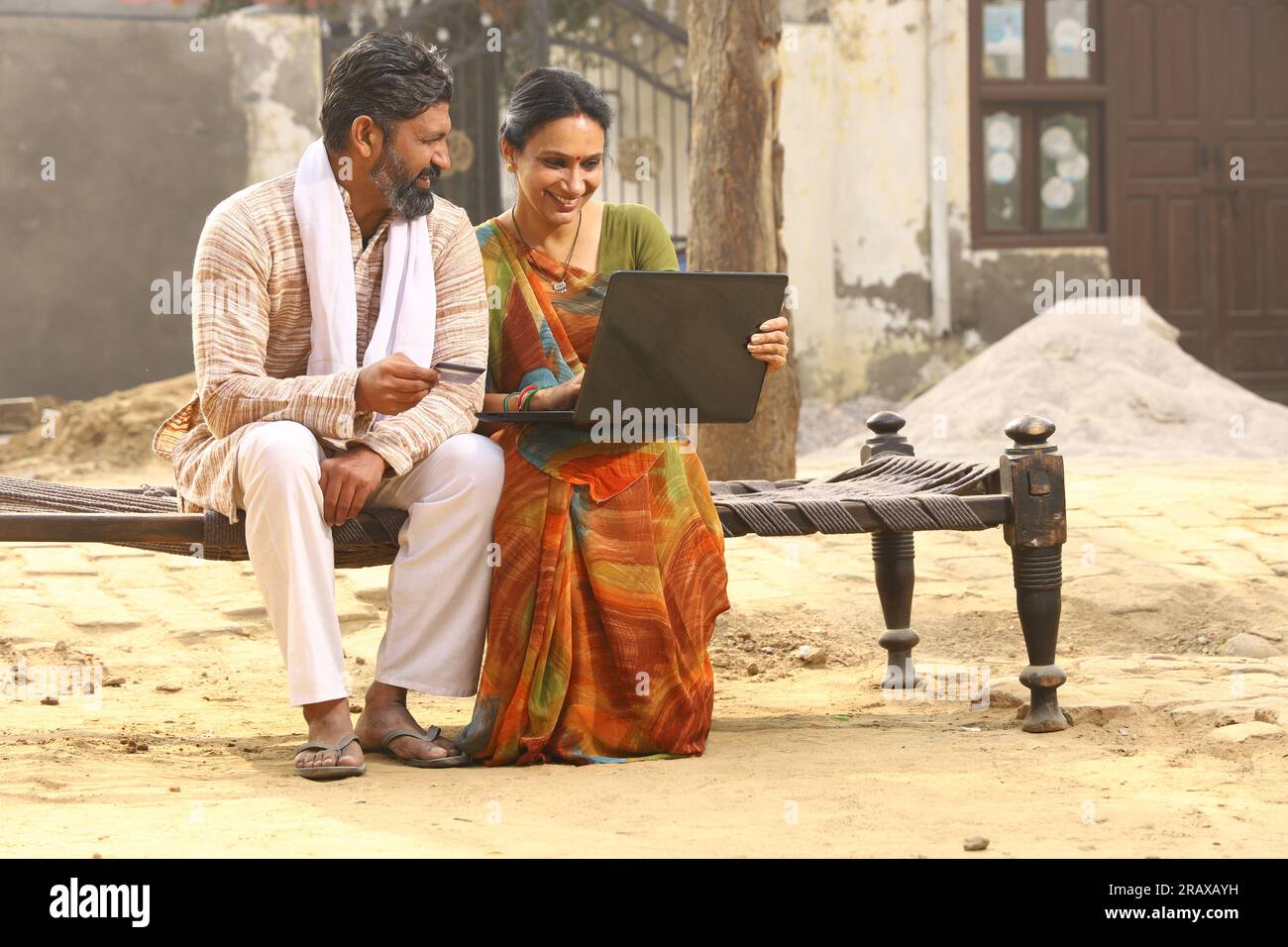 Heureuse famille indienne rurale dans le village. Couple rural assis ensemble souriant sur lit bébé devant leur maison dans la cour avant en utilisant un ordinateur portable pour les paiements en ligne. Banque D'Images