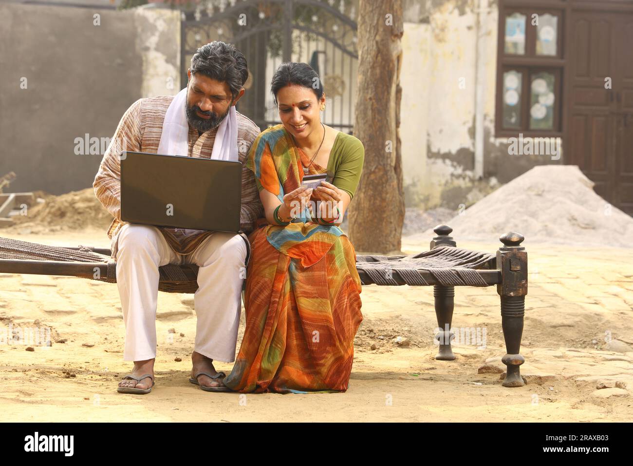 Heureuse famille indienne rurale dans le village. Couple rural assis ensemble souriant sur lit bébé devant leur maison dans la cour avant en utilisant un ordinateur portable pour les paiements en ligne. Banque D'Images