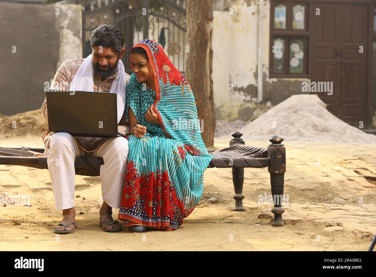 Heureuse famille indienne rurale dans le village. Mari et femme assis sur un lit bébé devant leur cour avant de la maison. homme en pyjama kurta et femme en belle saree. Banque D'Images