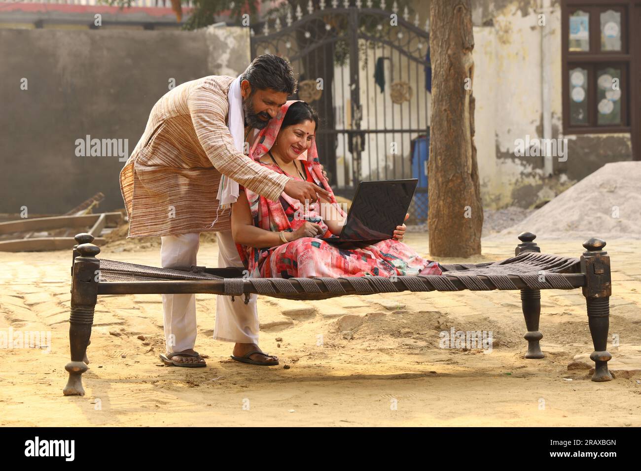 Heureuse famille indienne rurale dans le village. Mari et femme assis sur un lit bébé devant leur cour avant de la maison. homme en pyjama kurta et femme en belle saree. Banque D'Images