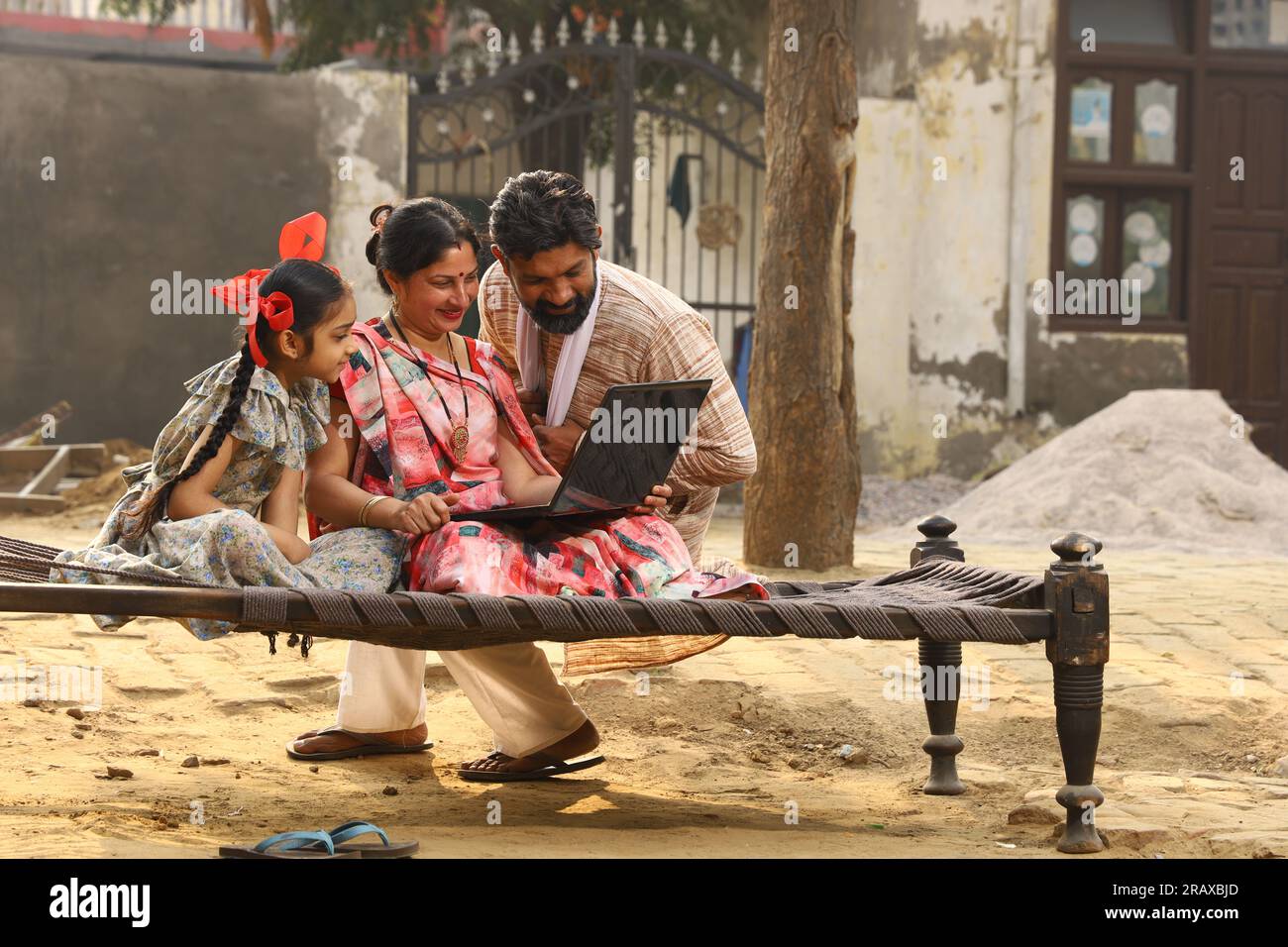 Happy Indian famille rurale mère, père, fille assis ensemble sur un lit à l'extérieur de leur cour avant à la maison en utilisant un ordinateur portable. Mère dans belle Saree. Indien Banque D'Images
