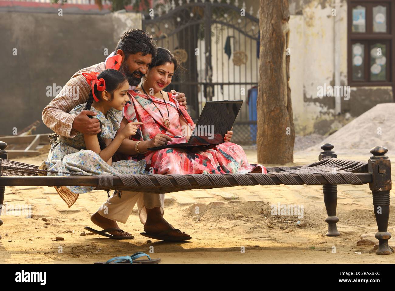 Happy Indian famille rurale mère, père, fille assis ensemble sur un lit à l'extérieur de leur cour avant à la maison en utilisant un ordinateur portable. Mère dans belle Saree. Indien Banque D'Images