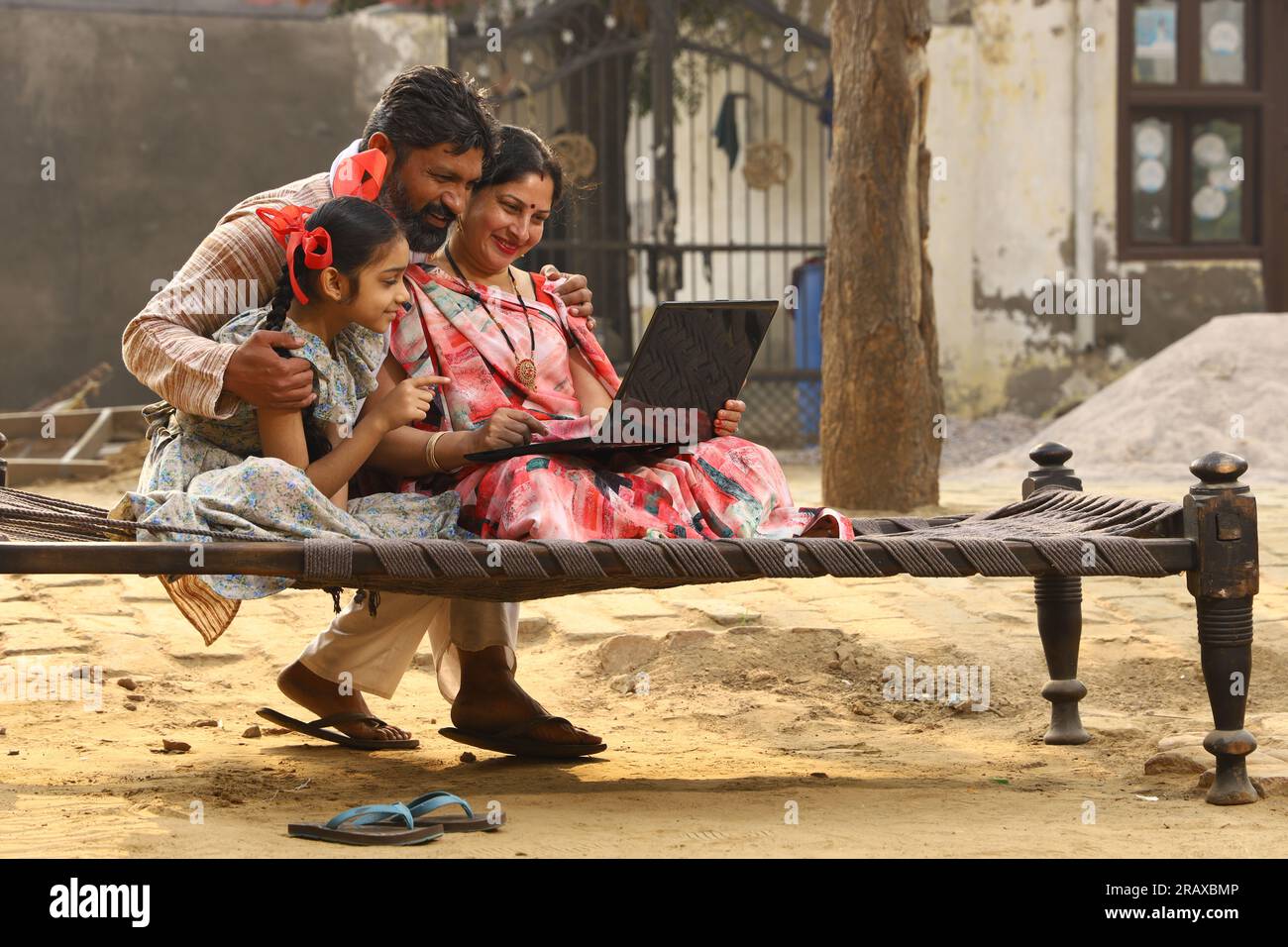 Happy Indian famille rurale mère, père, fille assis ensemble sur un lit à l'extérieur de leur cour avant à la maison en utilisant un ordinateur portable. Mère dans belle Saree. Indien Banque D'Images