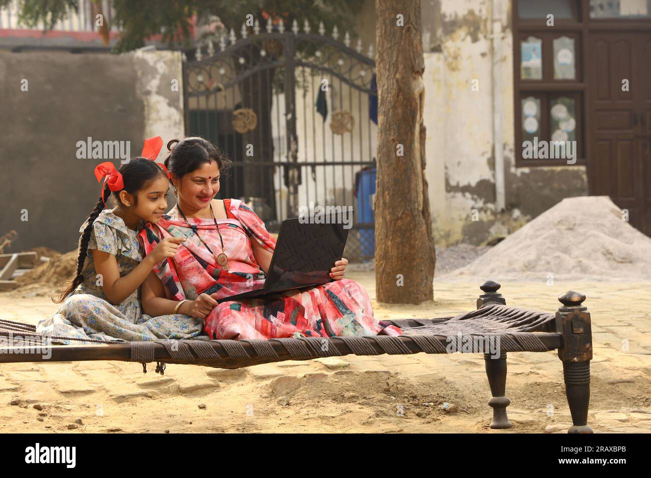 Happy Indian famille rurale mère, père, fille assis ensemble sur un lit à l'extérieur de leur cour avant à la maison en utilisant un ordinateur portable. Mère dans belle Saree. Indien Banque D'Images