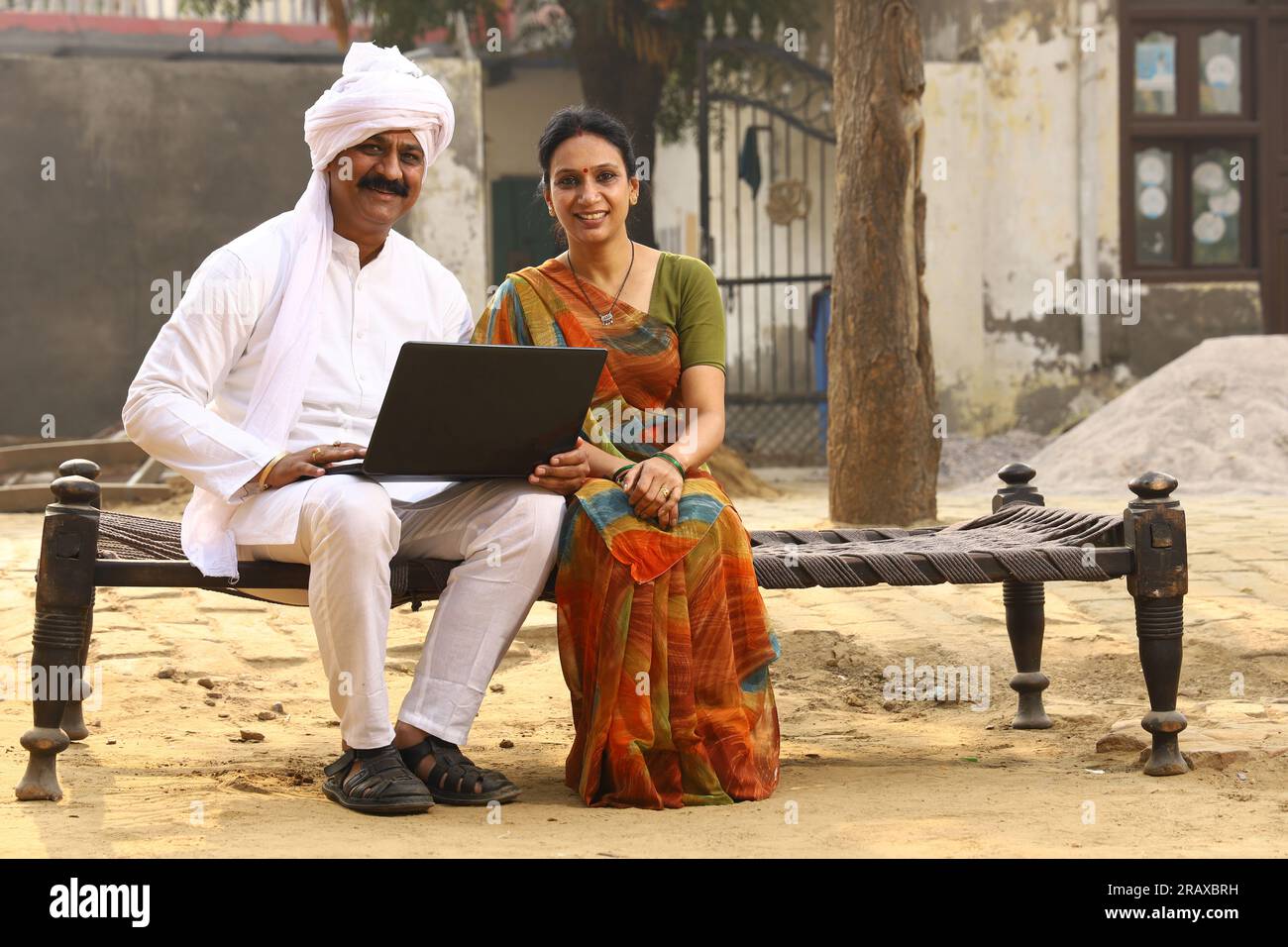 Heureuse famille indienne rurale dans le village. Mari et femme assis sur un lit bébé devant leur cour avant de la maison. homme en pyjama kurta et femme en belle saree. Banque D'Images