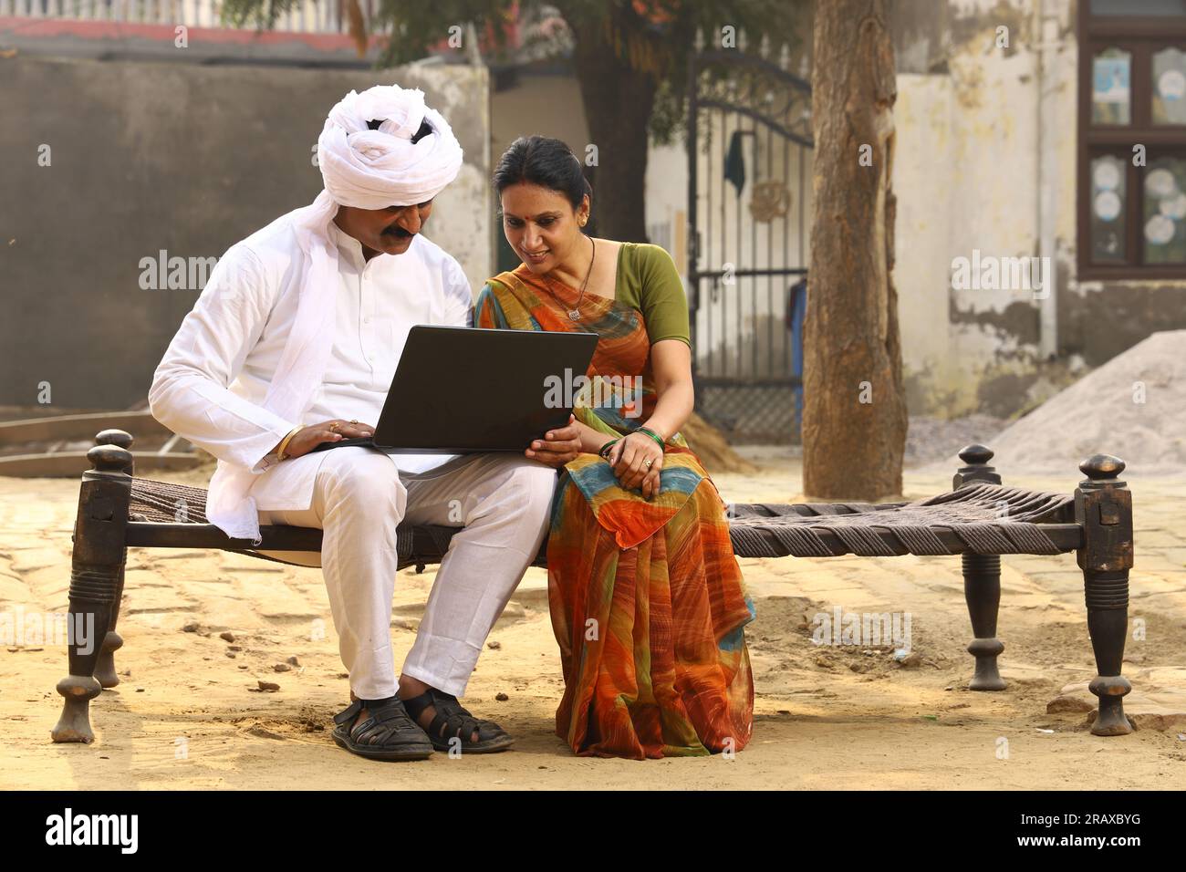 Heureuse famille indienne rurale dans le village. Mari et femme assis sur un lit bébé devant leur cour avant de la maison. homme en pyjama kurta et femme en belle saree. Banque D'Images