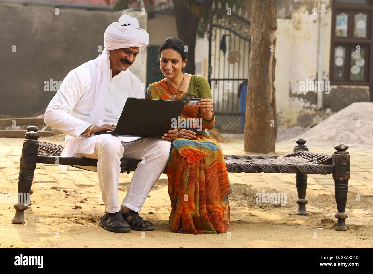 Heureuse famille indienne rurale dans le village. Mari et femme assis sur un lit bébé devant leur cour avant de la maison. homme en pyjama kurta et femme en belle saree. Banque D'Images