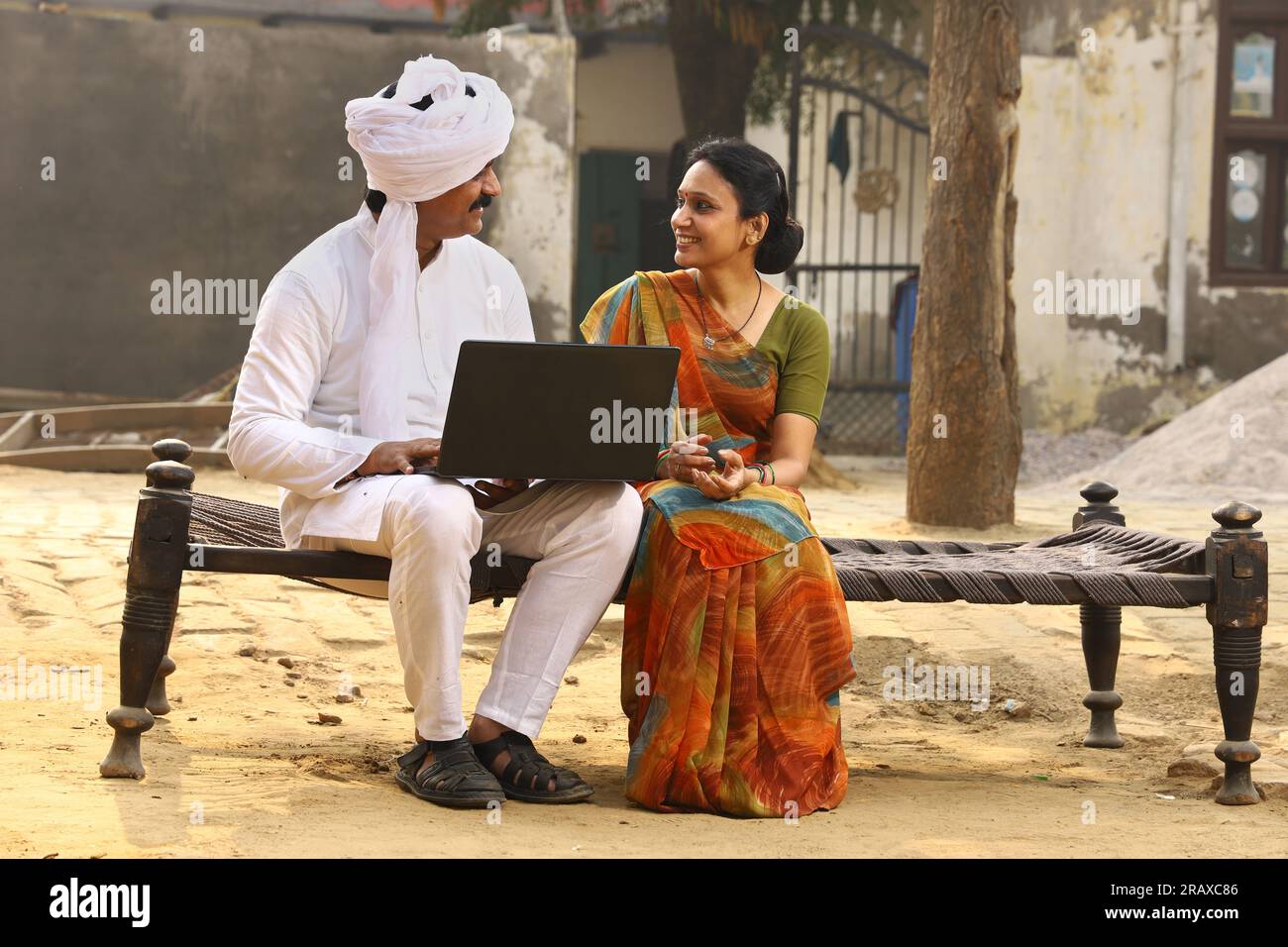 Heureuse famille indienne rurale dans le village. Mari et femme assis sur un lit bébé devant leur cour avant de la maison. homme en pyjama kurta et femme en belle saree. Banque D'Images