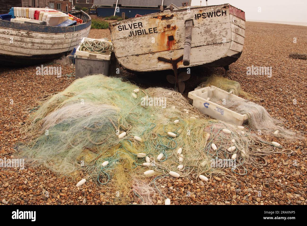 Deux petits vieux bateaux de pêche en bois sur la plage de galets à Aldeburgh, Suffolk. Royaume-Uni avec un tas de filets de chalutiers fins et des flotteurs couchés sur les pierres Banque D'Images