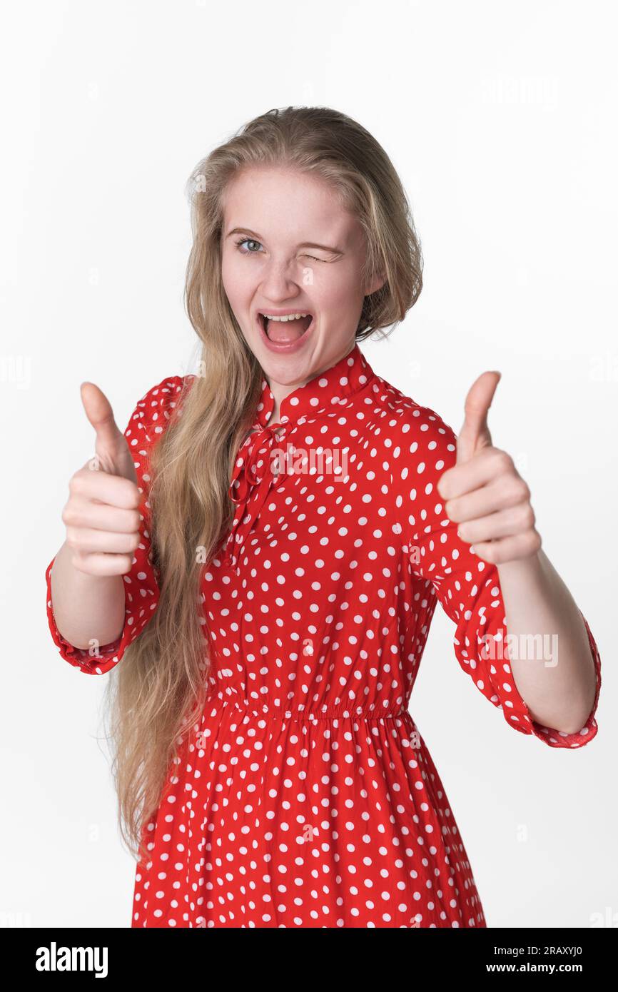 Bonheur blonde jeune femme ludique fermé un œil et montrant les pouces vers le haut avec les deux mains. Portrait de jeune femme adulte souriante habillée en été Banque D'Images