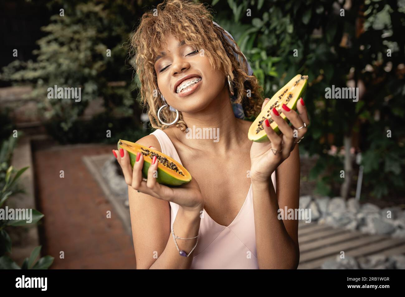 Souriant jeune femme afro-américaine avec bretelles waring tenue d'été et fermer les yeux tout en tenant la papaye fraîche dans le jardin intérieur flou, fashion-fo Banque D'Images