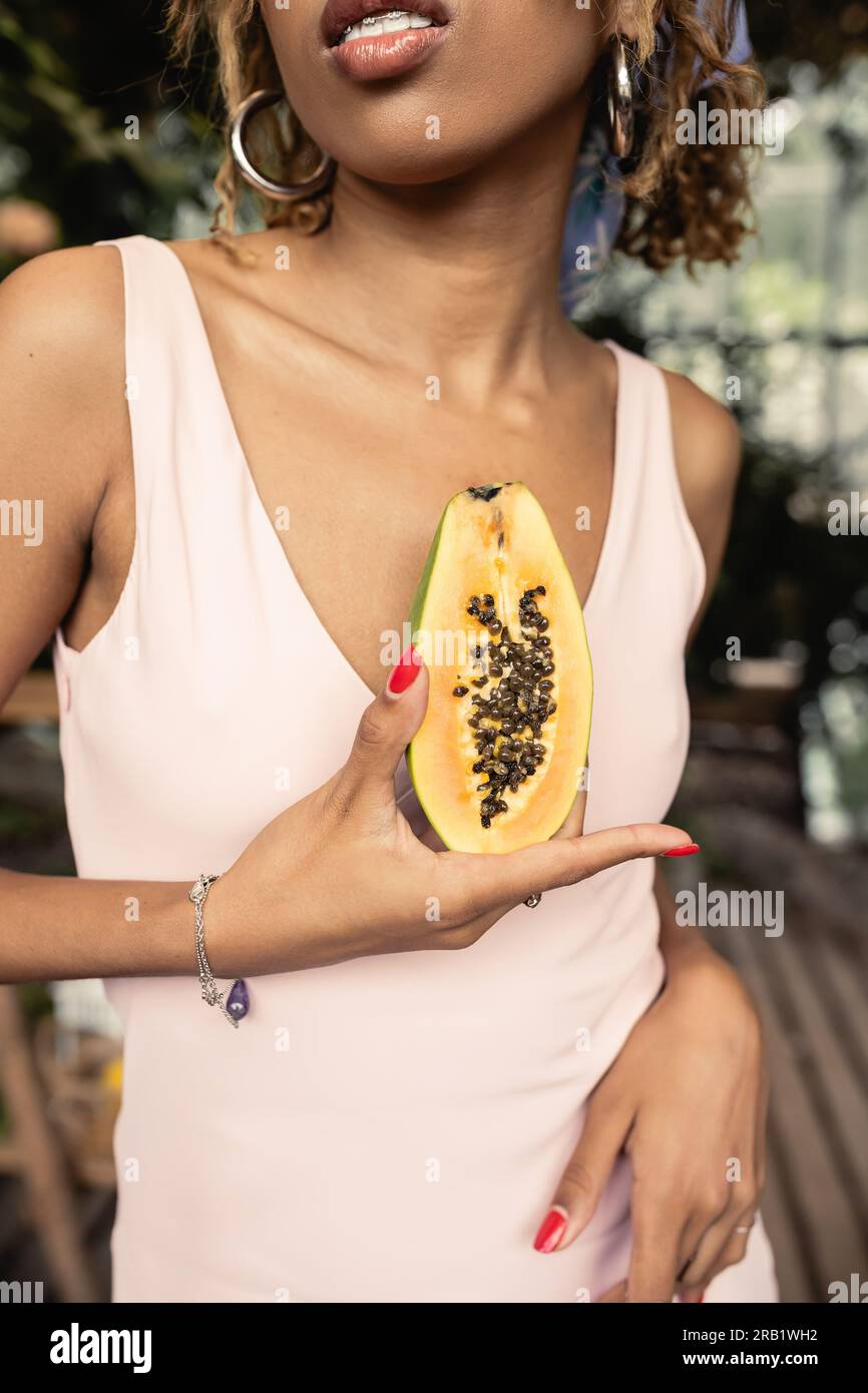 Vue courte de jeune femme afro-américaine élégante avec bretelles portant robe d'été tout en tenant la papaye mûre coupée dans le centre de jardin flou, mode- Banque D'Images