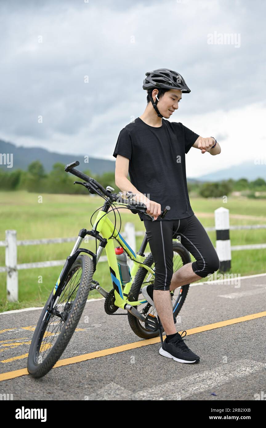 Un beau et heureux jeune homme asiatique en vêtements de sport et un casque de vélo se tient avec son vélo sur les routes de campagne et vérifie ses calories brûlées et ridi Banque D'Images