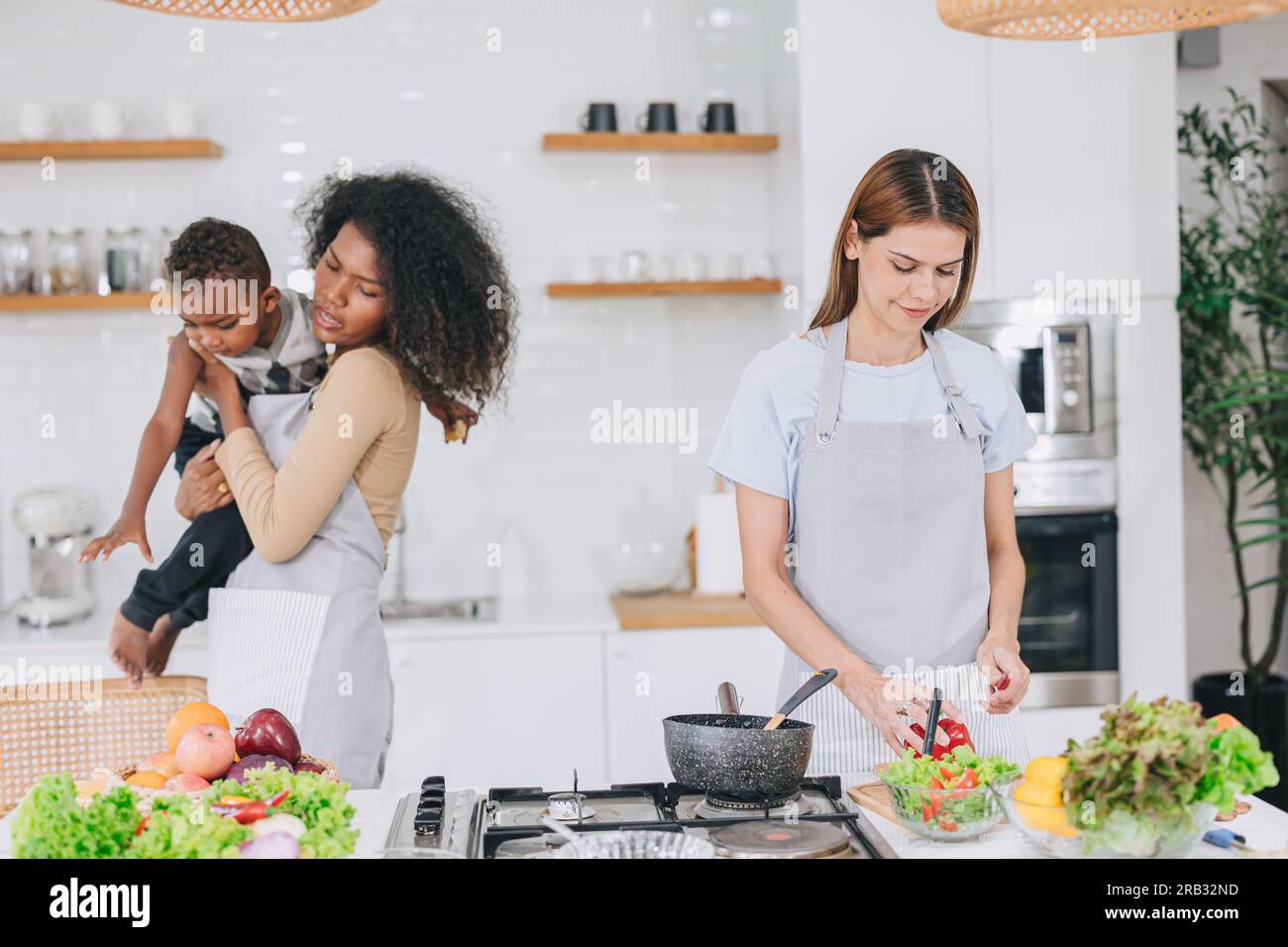 les femmes font la fête en cuisinant des repas à la maison de vacances de cuisine d'ami avec la garde d'enfant de fils ludique Banque D'Images