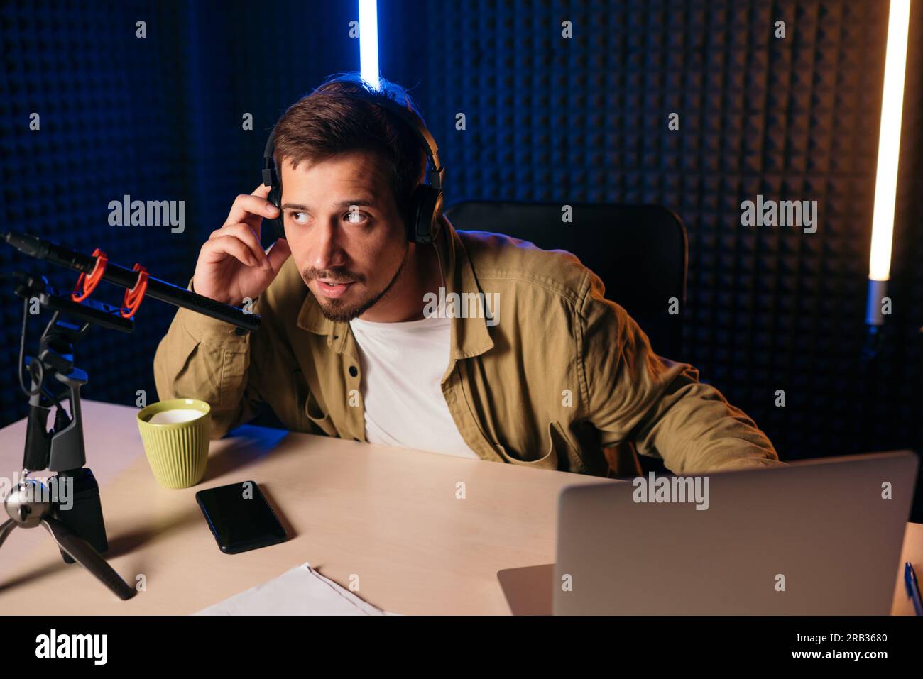 homme en chemise de moutarde avec des écouteurs faisant des gestes au microphone et partageant l'histoire avec le public tout en étant assis au bureau dans le studio avec éclairage au néon Banque D'Images