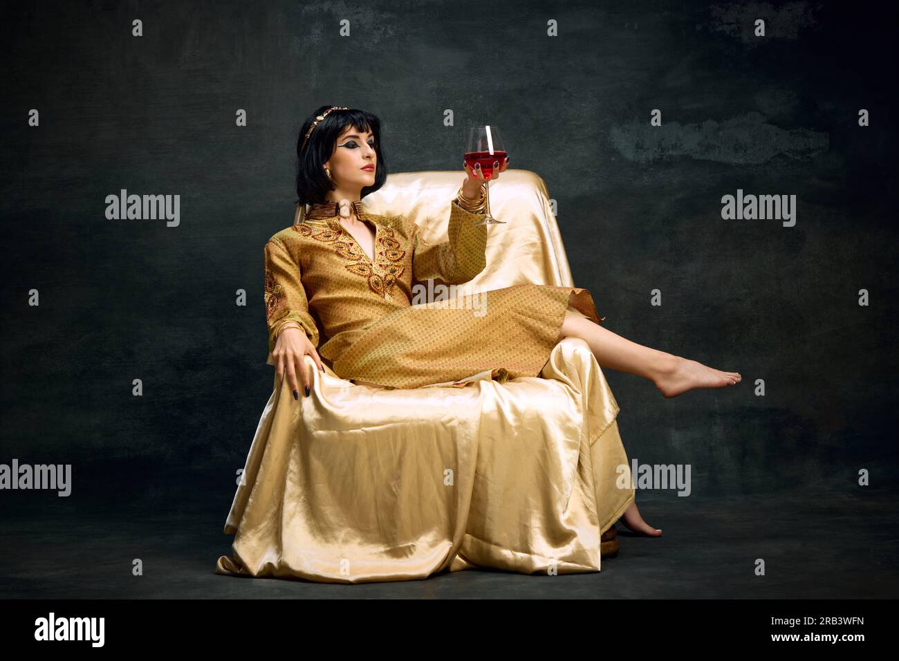 Portrait de noble, belle femme yougn en imag de reine, Cléopâtre, en robe goden assis et buvant du vin rouge sur fond vintage foncé Banque D'Images