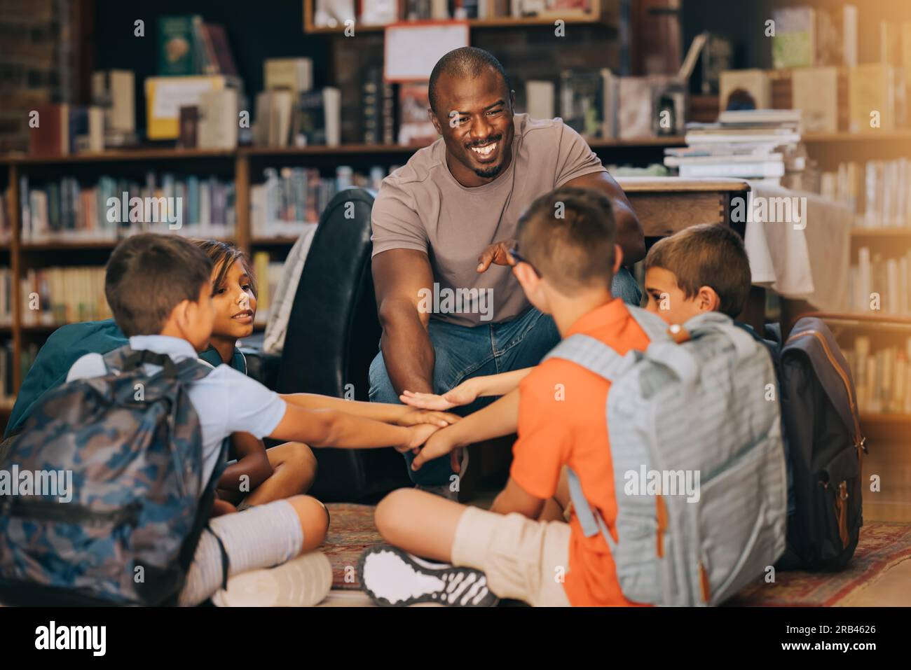Professeur d'école mâle se blottissant avec ses élèves dans une bibliothèque. Homme enseignant un groupe d'enfants de l'école primaire. Motivation et mentorat en primaire Banque D'Images