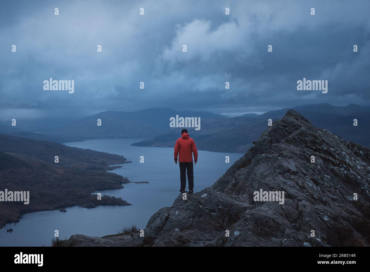 Un randonneur dans une veste rouge au sommet d'une montagne donne sur le paysage magnifique d'un lac entouré de montagnes. Loch Katrine. Loch Lomond et le parc national des Trossachs. Écosse Banque D'Images