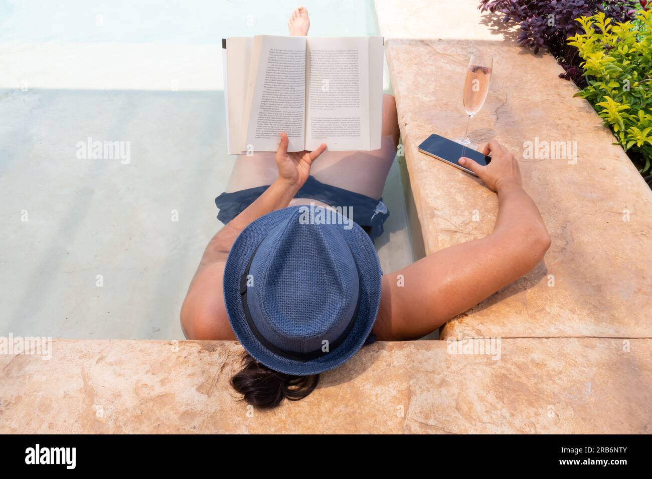 Photographié d'en haut et de derrière une femme dans un chapeau couché dans une piscine tenant un livre dans une main et un téléphone portable dans l'autre, à côté d'elle se trouve un verre o Banque D'Images